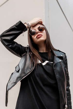 Stylish portrait of a young swag woman with fashionable trendy sunglasses in a stylish leather jacket and black sweatshirt near a gray wall on the street. Urban female rock look outfitの写真素材