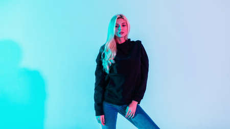 Beautiful fashionable girl model with blond hair in a stylish black hoodie and blue jeans in the studio on a bright neon pink backgroundの写真素材