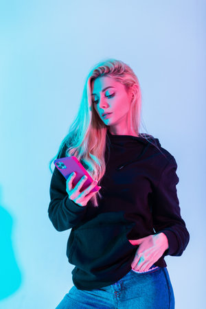 Fashion beautiful young woman in fashionable black hoodie with jeans uses smartphone in multicolored...