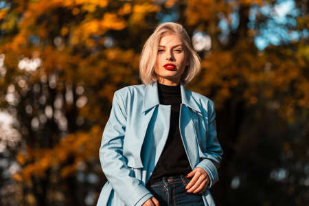 Elegant beautiful young blonde woman with red lips in a blue coat walks in an autumn park with orange fall foliage at sunsetの写真素材