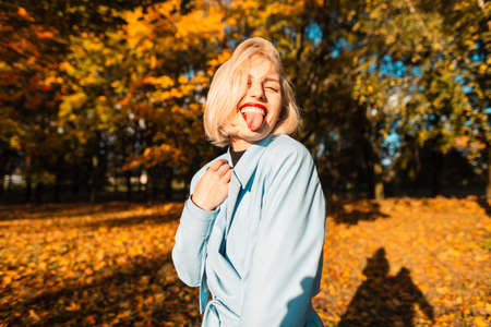 Funny beautiful young woman in fashionable clothes shows tongue and walks in autumn park at sunsetの写真素材