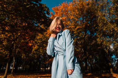 Happy portrait of a beautiful blonde girl with a smile in a fashionable blue coat relaxes outdoors in an autumn parkの写真素材