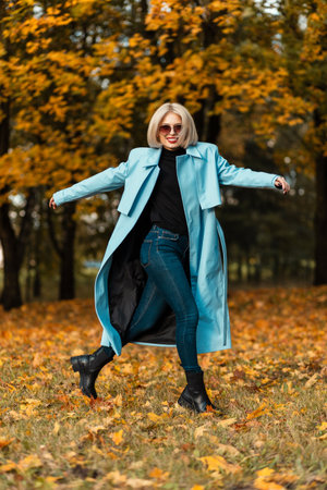 Happy beautiful young smiling girl in a fashionable blue coat with a sweater and jeans motion walks in an autumn park with colorful bright foliageの写真素材