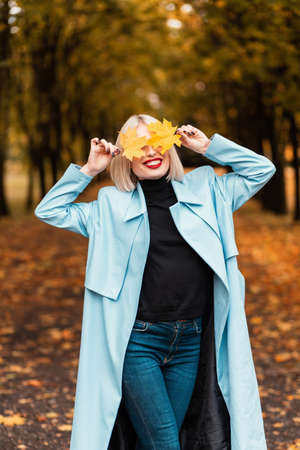 Funny smiling beautiful girl in fashionable bright clothes covers her face with yellow foliage and walks in the autumn parkの写真素材