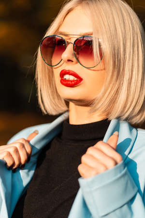 Fall portrait of a beautiful young stylish woman with red lips in fashionable clothes with vintage stylish sunglasses on nature at sunsetの写真素材