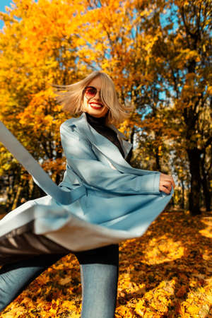 Happy young smiling girl in fashionable sunglasses with a bright blue coat walks motions in a park with yellow autumn foliage. Funny woman outdoors on sunny dayの写真素材