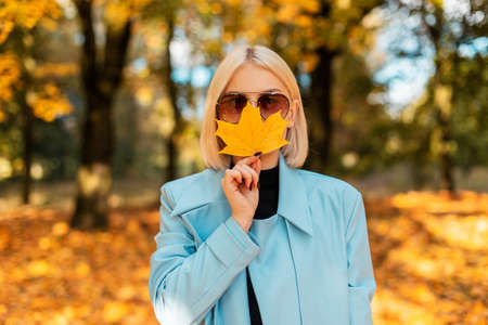 Creative portrait of a beautiful young girl with sunglasses in a fashion blue coat covering her lips with an autumn yellow leaf in a Canadian park with fall golden foliage. Amazing sunny vacationの写真素材