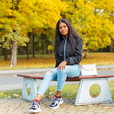 Beautiful young african american woman in fashionable clothes sitting on a park with yellow autumn foliageの写真素材