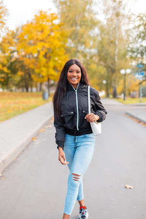 Pretty smiling black girl with trendy casual clothes with a fashion jacket, jeans and a stylish handbag is walking in an autumn park with bright yellow fall foliageの写真素材