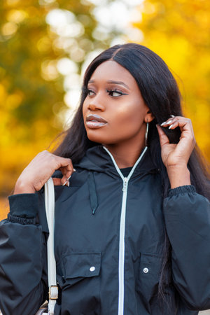Fashion portrait of a beautiful young African American woman in a stylish black jacket with a handbag straightens her hair and walks in the park against a background of colorful yellow autumn foliageの写真素材
