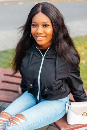 Portrait of a beautiful young black woman with a smile in a fashionable casual jacket with blue jeans and a bag sits on a park benchの写真素材