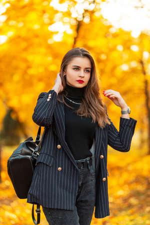 Elegant beautiful young woman model with red lips in a fashionable black blazer and sweater with a leather backpack walks in the park with yellow autumn foliage. Business female style outdoorsの写真素材