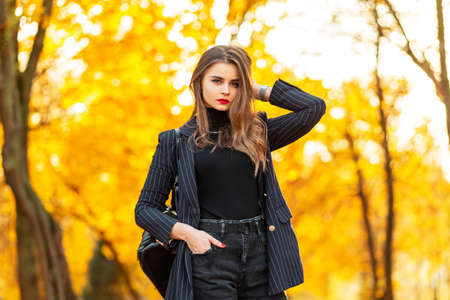 Beautiful business woman with red lips in a fashionable elegant suit with a blazer, sweater and backpack walks in the nature with bright colored golden autumn foliageの写真素材