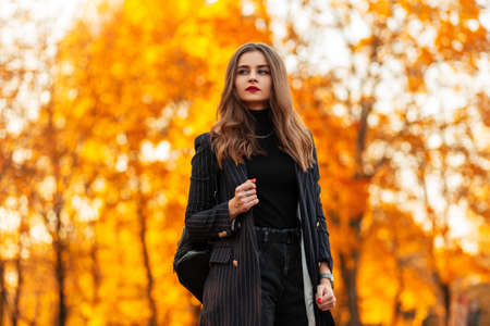 Beautiful molded fashionable girl with red lips in a fashion clothes with a blazer and a sweater with a leather backpack walks in the park with orange leaves at sunsetの写真素材