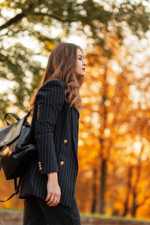 pretty elegant woman in fashionable black suit with stylish blazer and leather backpack walks in the park with colored yellow autumn foliage at sunsetの写真素材