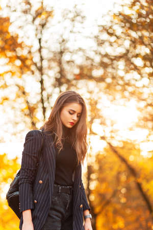 beautiful girl in a trendy business suit with a stylish blazer and a sweater with a fashion backpack walks in the park with golden autumn foliage at sunsetの写真素材