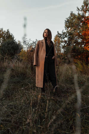 Beautiful fashion young woman in a beige coat, black sweater and jeans on a nature in a field with grassの写真素材