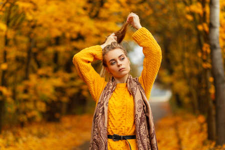Beautiful girl in a fashionable knitted yellow sweater with a style scarf walks in the park with amazing yellow autumn foliageの写真素材
