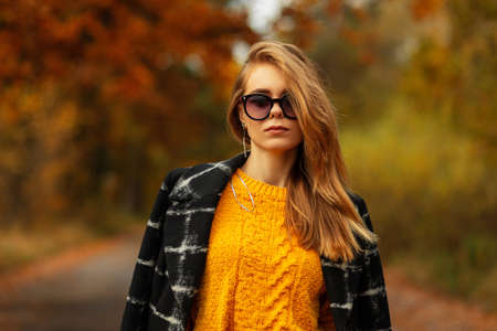 Portrait of a stylish caucasian girl with neutral emotions with fashionable sunglasses in an autumn yellow sweater and black coat walks in a park with orange autumn foliage, outdoorsの写真素材
