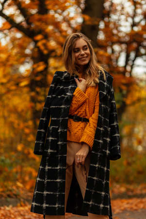 Happy fashionable young woman with a pretty smile in trendy clothes look with a coat and a sweater walks in an autumn park with colorful bright foliageの写真素材
