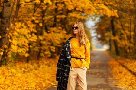 Stylish beautiful young woman with sunglasses in fashion autumn clothes look with a knitted sweater and black coat against the background of an amazing autumn park with yellow foliage. girl outdoorsの写真素材