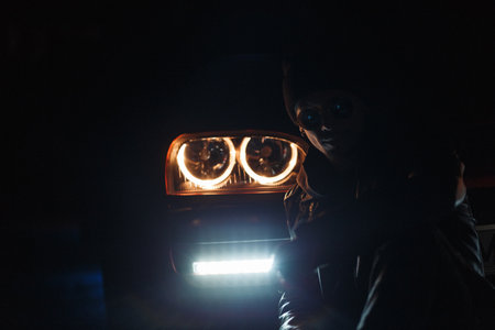 Stylish young man in fashionable sunglasses with a leather jacket and a hat sits in the dark near a car with bright headlightsの写真素材
