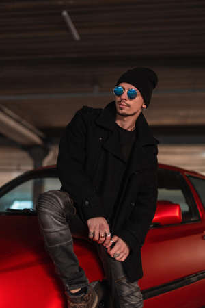 Elegant casual handsome man in stylish blue sunglasses in a black fashionable coat with a hat sits...