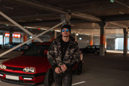 Fashionable handsome man model with cool sunglasses in a stylish military jacket walks near a classic red car at the parkingの写真素材