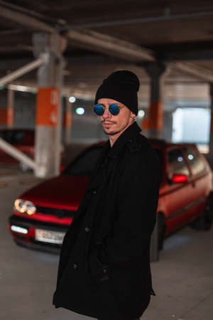 Young fashion man with stylish blue sunglasses in vintage coat and hat near a old red carの写真素材