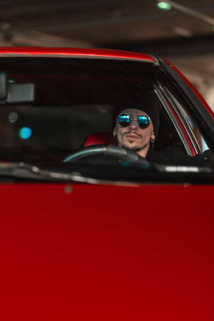 Stylish young man in sunglasses and a hat sits at the wheel of an old red car. male taxi driver drivingの写真素材