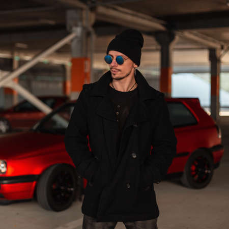 Handsome young man with sunglasses in fashionable black coat with hat near red car in cityの写真素材