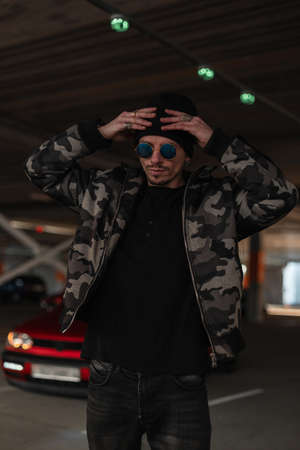 Handsome young man model in stylish sunglasses in fashionable clothes with winter military jacket wear a black hat in cityの写真素材