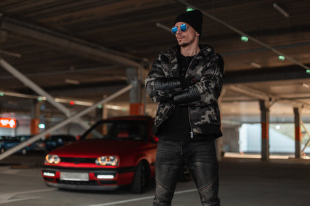 Cool young man model with stylish sunglasses and black hat in fashion winter military jacket walks on the streetの写真素材