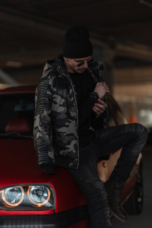 Cool hipster guy in street clothes with a military jacket, hat, and glasses stands in front of his car in the parking lotの写真素材