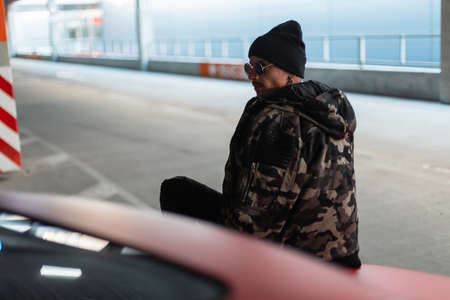 Stylish handsome young hipster man with sunglasses and a black hat in a fashionable winter military jacket sits on a red car in the cityの写真素材