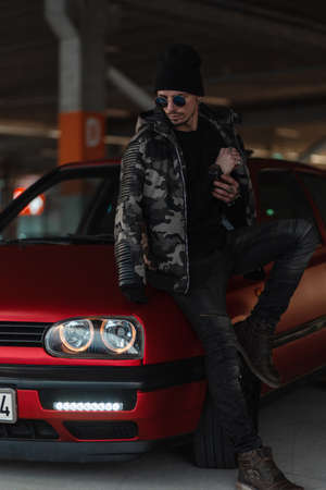 Fashionable handsome man model with sunglasses and hat in stylish winter military jacket with pullover and jeans near red car on the streetの写真素材