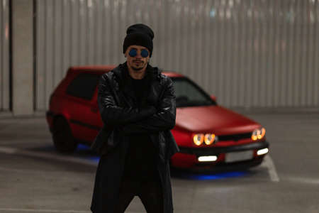 Cool stylish guy with fancy black glasses and a hat in a leather jacket and hoodie stands in a...