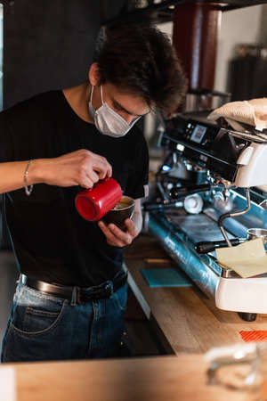 Young guy barista with medical mask makes delicious cappuccino coffee with coffee in a cafe. Covid-19 and pandemic protection conceptの写真素材