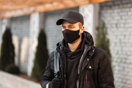 Young man with a black protective mask and a fashionable cap in a black stylish hoodie and a winter...