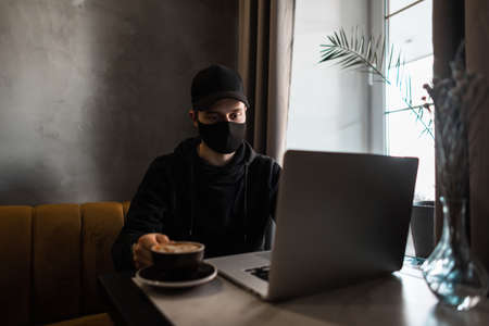 Young guy with a medical protective mask and a black cap in a hoodie drinks coffee and looks at a...