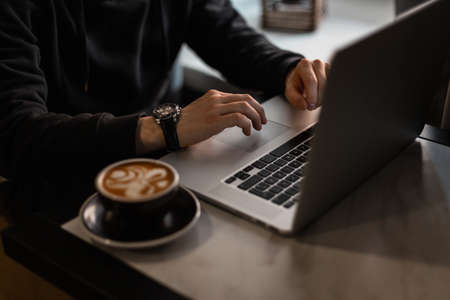 Professional man with luxury watch in black hoodie works on laptop and drinks coffee indoors