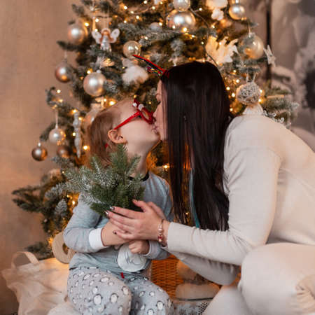 Beautiful happy mother with her little daughter makes a kiss on the background of a Christmas tree and lights. Winter family holidays at homeの写真素材
