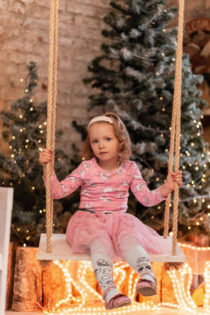 Beautiful little girl child in a fashionable pink dress sits and rolls on a swing against the background of Christmas trees and lights at homeの写真素材
