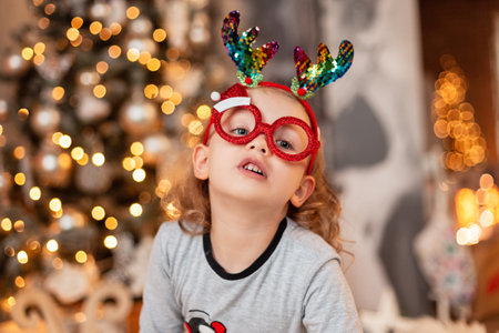 Funny little girl with fun red Santa glasses and deer horns in fashionable pajamas against the...