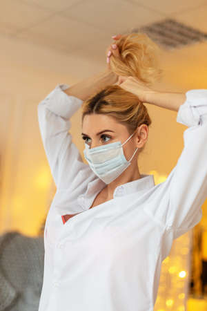 Beautiful young woman doctor with a medical mask in a white shirt straightens her hair indoors with yellow lightsの写真素材