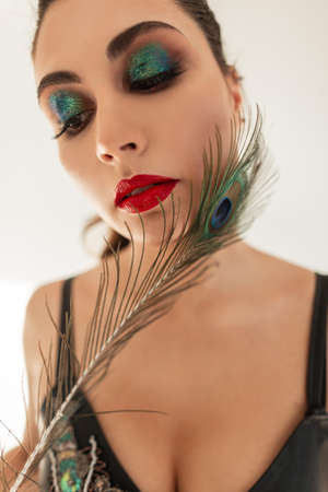 Pretty young stylish glamorous woman with professional makeup, red lips and green eyelids holding a peacock feather near her faceの写真素材