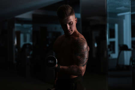 Cool strong young man fitness trainer with a hairstyle and a naked torso with muscles doing hard exercises and workout biceps with a dumbbell in the gym at night.の写真素材