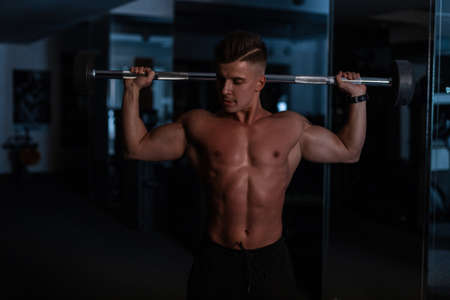 Strong handsome guy with a naked muscular body holding the bar and working out in the gym on a dark backgroundの写真素材