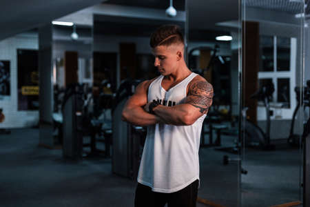 Young handsome man athlete with hairstyle in a white t-shirt posing in the sport gym on a dark...