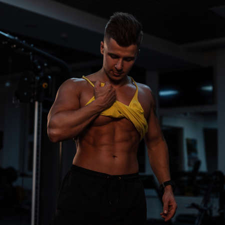 Sexy muscular guy athlete with a tanned body lifted a T-shirt and shows abdominal muscles on a dark background. Bodybuilder training in the gymの写真素材